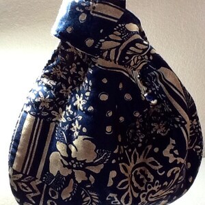 May include: A navy blue and beige patterned fabric bag with a knotted handle. The bag features a floral and geometric design. The fabric has a soft, textured appearance. The bag is likely handmade and suitable for carrying small items.