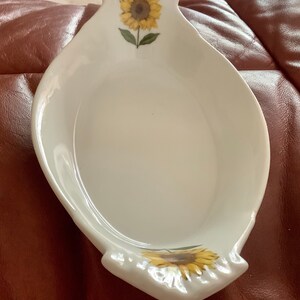 May include: White ceramic spoon rest with a sunflower design. The spoon rest has a curved shape with a sunflower painted at the top and bottom. The sunflower has yellow petals and a brown center.