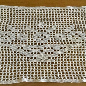 May include: White crocheted rectangular doily featuring an openwork heart design in the center. The doily is made of a fine, delicate yarn, and the open weave creates a lacy effect. The background is a light brown wooden surface.