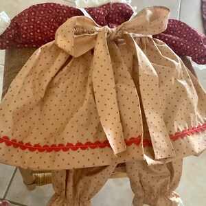 Op de afbeelding: Een baby-outfit met een tan en rood patroon en een grote strik. De outfit bevat een top met een strik, een broek en een bijpassende muts. De outfit heeft een rode geschulpte rand en is gemaakt van een zachte stof.