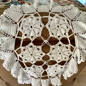 May include: A white, handmade crochet doily with a floral design. The doily features a central pattern of stylized flowers and an outer scalloped edge. The intricate openwork design is visible throughout the piece, showcasing the craftsmanship.