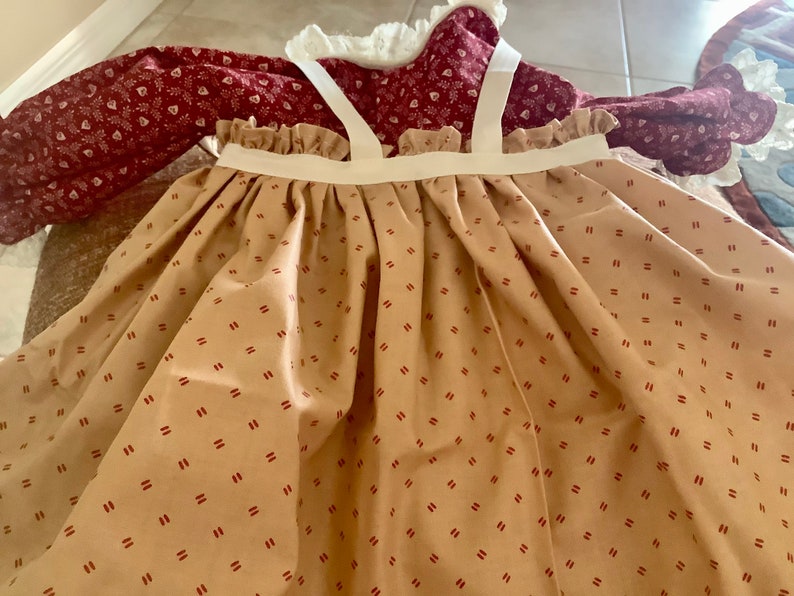 Op de afbeelding: Een jurk in vintage-stijl met een beige rok met een herhalend patroon van kleine rode streepjes. De lijfje is dieprood met een bloemenpatroon en de jurk heeft witte bandjes en een gerimpelde halslijn. De jurk lijkt van katoen te zijn.