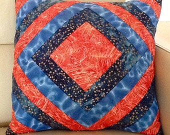 May include: Square decorative pillow with a quilted design. The pillow features a central red square, surrounded by blue and navy blue bands with a star pattern. The pillow is placed on a neutral-colored surface.