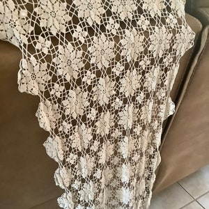 May include: Cream-colored lace table runner with a floral pattern. The delicate openwork design features repeating flower motifs connected by a web-like structure. The edge of the runner has a scalloped trim.