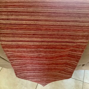 May include: A rectangular table runner with a red and beige horizontal striped pattern. The runner is made of a woven fabric and has a pointed end. The runner is draped over the arm of a beige chair.