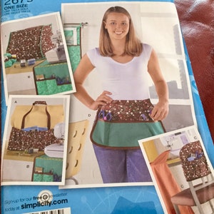 May include: Simplicity sewing pattern 2679. The pattern features a brown, green, and tan apron with pockets, decorated with pins and scissors. The pattern also includes a sewing machine cover and a tote bag. The model is wearing a white top and jeans.