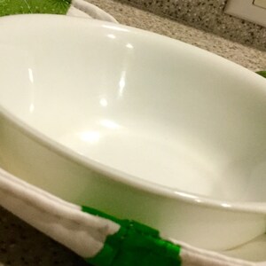 May include: A white oval-shaped bowl rests on a green and white patterned fabric. The bowl is made of a glossy, reflective material. The fabric appears to be a quilted potholder or bowl cozy, providing insulation. The background is out of focus.