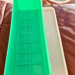 May include: A green plastic ice cube tray with a rectangular shape and a grid pattern. A white plastic lid is next to the tray. The tray is empty and ready to be filled with water.