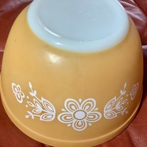 May include: An upside-down, vintage, yellow mixing bowl with a white floral design. The bowl has a rounded shape and a white interior. The floral design includes stylized flowers and leaves. The bowl is made of glass and is likely from the mid-20th century.