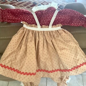 Op de afbeelding: Een baby-outfit in vintage-stijl met een bordeauxrood topje met bloemenprint, witte kanten rand en lange mouwen. De outfit bevat een beige rok met een geschulpte rode zoom en bretels. De outfit is gemaakt van zachte stof.