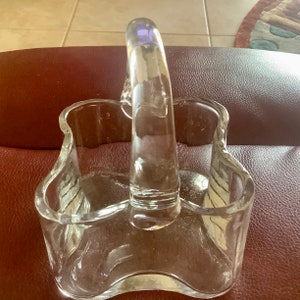 May include: Clear glass basket with a curved handle. The basket has a wavy rim and a rectangular shape. The handle is arched and attached to the sides of the basket. The glass is transparent, allowing light to pass through.