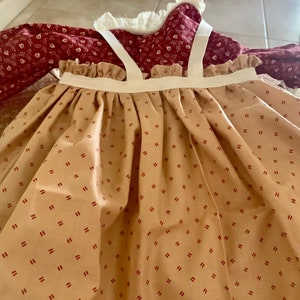 Op de afbeelding: Een vintage jurk met een beige rok met een patroon van kleine rode streepjes. De lijfje is dieprood met een bloemenpatroon en de jurk heeft witte bandjes en een gerimpelde halslijn. De jurk lijkt handgemaakt te zijn.
