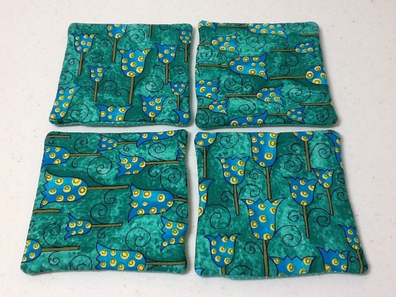 Puede incluir: Cuatro posavasos cuadrados de tela con un fondo verde azulado y un patr&oacute;n repetido de flores estilizadas azules y amarillas. Los posavasos est&aacute;n hechos de una tela suave y texturizada.