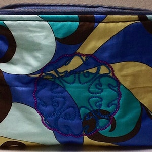May include: A rectangular, quilted cosmetic bag with a blue zipper and a small ribbon pull. The bag features a colorful abstract pattern in shades of blue, teal, yellow, and brown. An embroidered design with a pink outline is centered on the front.