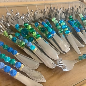 May include: A collection of stainless steel forks with handles adorned with blue, green, and clear glass beads. The beads are wrapped with gold-colored wire, creating a decorative and unique look. The forks are arranged on a reflective surface.