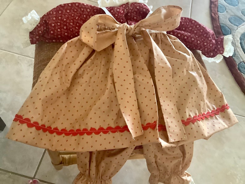 Op de afbeelding: Een baby-outfit met een tan en rood patroon en een grote strik. De outfit bevat een top met een strik, een broek en een bijpassende muts. De outfit heeft een rode geschulpte rand en is gemaakt van een zachte stof.