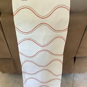 May include: White fabric runner with a wavy pattern of red and gray dots. The runner is draped over a brown surface, showcasing its design. This decorative piece adds a touch of elegance to any table setting.