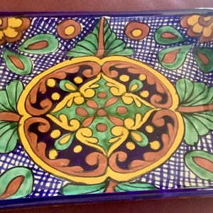 May include: Rectangular ceramic serving dish with a vibrant hand-painted design. The dish features a deep blue border and a central pattern of green, yellow, and brown floral and leaf motifs. The background has a white grid pattern.