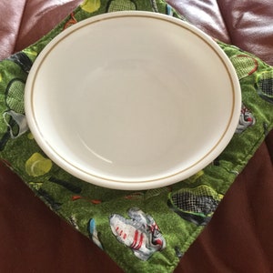 May include: A white ceramic bowl with a gold rim sits on a green, square quilted hot pad. The hot pad features a tennis theme with tennis rackets, balls, and shoes. The hot pad is approximately 8 inches square.