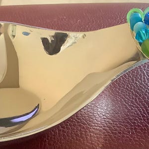 May include: A silver-colored spoon rest with a decorative beaded handle. The handle is adorned with an array of blue and green beads. The spoon rest is made of a shiny, reflective metal.