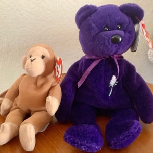 May include: Two plush toys: a brown monkey and a purple bear. The bear has a ribbon around its neck and a flower embroidered on its chest. Both toys have tags attached.
