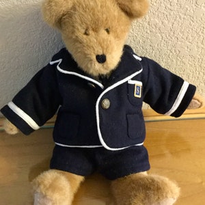 May include: A plush teddy bear wearing a navy blue uniform with white trim. The uniform includes a jacket with a gold button and a small patch with the letter "S", and matching shorts. The bear has light brown fur.