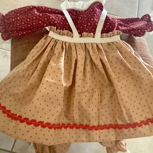 Op de afbeelding: Een vintage jurk met een bordeauxrood lijfje met bloemenprint en een beige rok versierd met kleine bruine streepjes. De rok heeft een geschulpte rode rand. Witte bandjes en kanten details maken dit klassieke kledingstuk compleet.