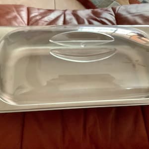 May include: A stainless steel food pan with a clear, curved lid. The lid has two raised handles for easy opening. The pan is rectangular and appears to be empty. The image is taken indoors.