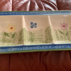 May include: Rectangular ceramic serving dish with three compartments. The dish features a white interior with painted flowers and a butterfly. A blue and green border with a decorative swirl design frames the dish.
