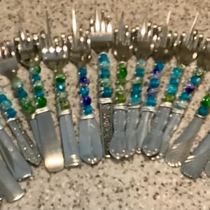 May include: A collection of silver forks with light blue handles. The handles are decorated with a string of blue and green beads. The forks are arranged in a semi-circle on a speckled surface.