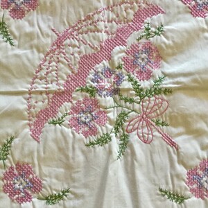 May include: A white fabric with a cross-stitch design featuring a pink parasol, pink and purple flowers, and green foliage. A pink bow is also part of the design. The embroidery is detailed and the overall aesthetic is vintage.