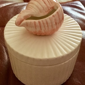 May include: A white ceramic trinket box with a ridged lid, topped with a pink seashell-shaped container. The seashell has vertical stripes and a small opening. The box and seashell are on a brown surface.