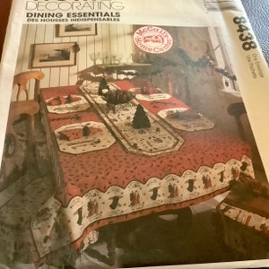 May include: A red tablecloth with a white scalloped border featuring figures and text. The tablecloth is set with matching placemats and a runner. The image also shows a chair and a framed picture on the wall. The text on the image says "Dining Essentials".