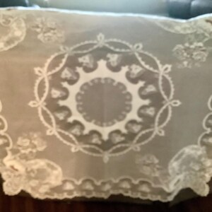 May include: A sheer, white lace tablecloth with a floral and circular design. The edges feature a scalloped trim. The delicate fabric is draped over a brown surface, creating a soft, elegant look. This vintage-style table covering is suitable for special occasions.