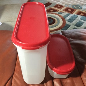 May include: Two white plastic food storage containers with red lids. The larger container is tall and rectangular, while the smaller one is oval. Both containers are empty and ready to be filled with food.