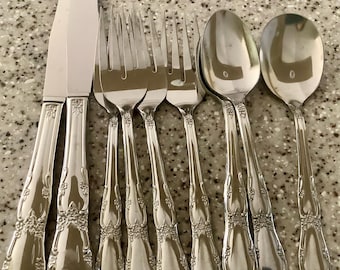 May include: A collection of silver-colored flatware, including a butter knife, forks, spoons, and a serving spoon. The handles feature a floral design. The flatware is arranged on a speckled surface.