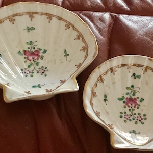 May include: Two white, shell-shaped porcelain dishes with floral designs. Each dish features a pink rose and green leaves in the center, with a gold trim and a decorative border. The dishes are on a brown surface.