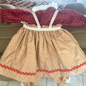 Op de afbeelding: Een baby-outfit in vintage-stijl. Het bordeauxrode lijfje heeft een bloemenprint, witte kanten randen en lange mouwen. De beige rok heeft een rode geschulpte zoom en bretels. Gemaakt van zachte stof.
