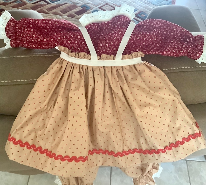 Op de afbeelding: Een baby-outfit in vintage-stijl. Het bordeauxrode lijfje heeft een bloemenprint, witte kanten randen en lange mouwen. De beige rok heeft een rode geschulpte zoom en bretels. Gemaakt van zachte stof.