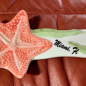 May include: Ceramic starfish-shaped spoon rest with a coral-colored starfish design. The white handle features the text "Miami, FL" in black script, along with green and coral accents. A decorative kitchen item.