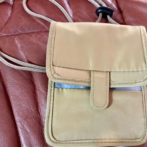 May include: A tan nylon neck wallet with a matching cord and black plastic cord lock. The wallet has a flap closure and a small pocket on the front. The stitching is a matching tan color.