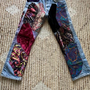 Pode incluir: Jeans de ganga clara com detalhes de patchwork. Retalhos de veludo e tecidos estampados em tons de vermelho, roxo e dourado são costurados na frente. Os jeans têm braguilha com botões e design clássico de cinco bolsos.