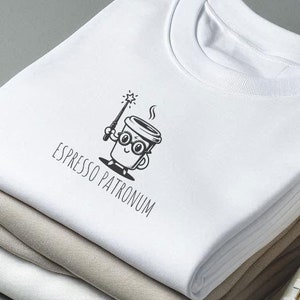 Könnte beinhalten: Ein weißes T-Shirt mit einer schwarz-weißen Illustration einer Kaffeetasse mit einem Zauberstab und dem Text "Espresso Patronum". Das T-Shirt ist gefaltet und mit anderen weißen und beigefarbenen T-Shirts gestapelt. Zwei weiße Canvas-Sneaker befinden sich im Vordergrund.