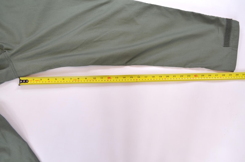 May include: An olive green garment sleeve with a yellow measuring tape laid across it. The tape measure shows a length of approximately 76 cm. The sleeve has a velcro patch at the cuff.