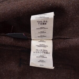 May include: Close-up of a brown wool fabric with two attached white tags. The tags have text in multiple languages, including Japanese. The fabric appears to be part of a garment.
