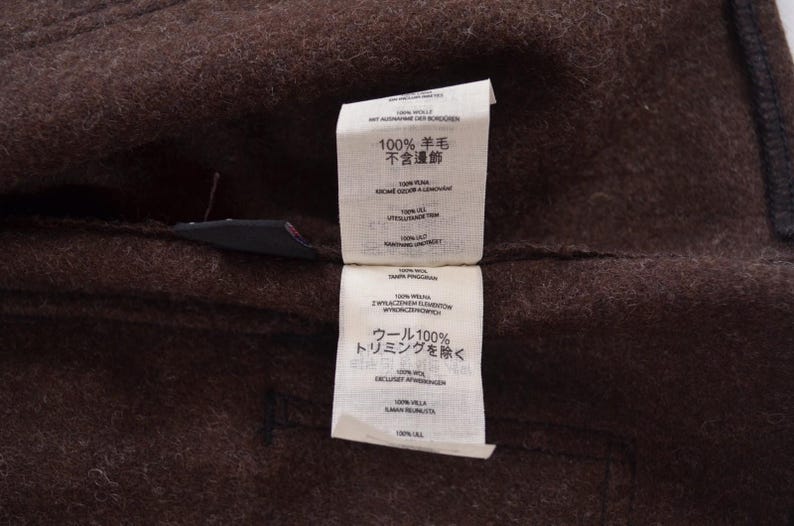 May include: Close-up of a brown wool fabric with two attached white tags. The tags have text in multiple languages, including Japanese. The fabric appears to be part of a garment.