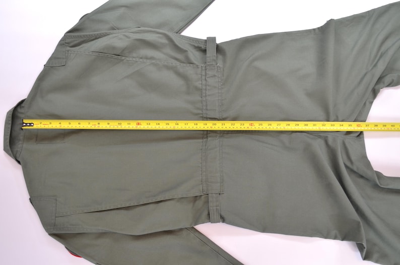 May include: Olive green jumpsuit with a yellow measuring tape laid across the front. The jumpsuit has a collar, pockets, and a belt loop. The measuring tape shows a length of approximately 76 cm.