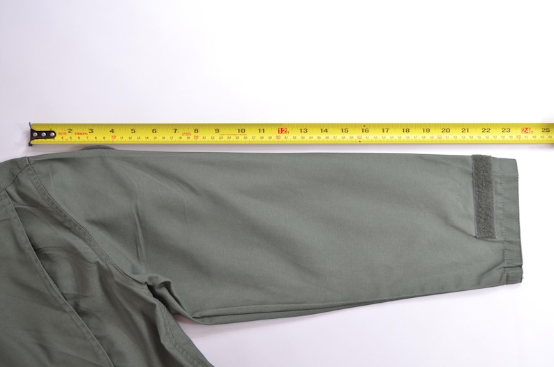 May include: A close-up of a sage green jacket sleeve with a yellow measuring tape laid across it. The sleeve has a velcro closure at the cuff. The measuring tape shows a length of approximately 61 cm.
