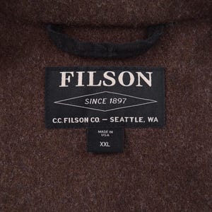 May include: A close-up of a brown wool coat with a black collar. A black label reads "FILSON SINCE 1897" and "C.C. FILSON CO. - SEATTLE, WA". A smaller label indicates "MADE IN USA" and size "XXL".