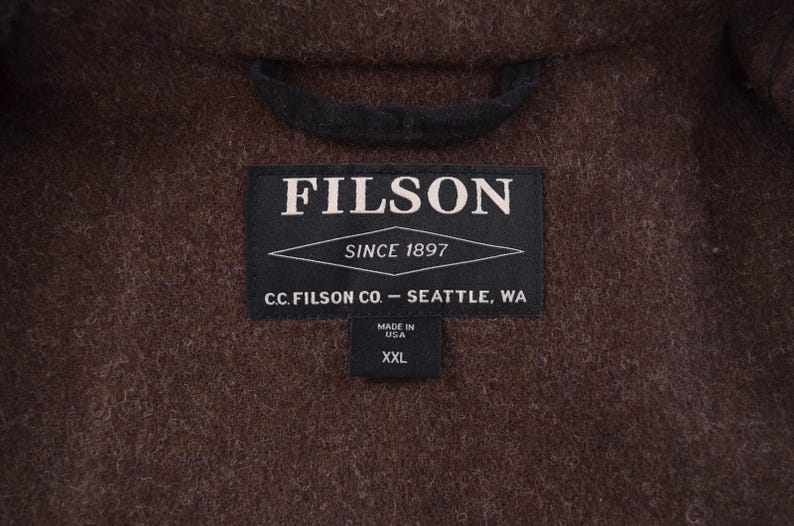 May include: A close-up of a brown wool coat with a black collar. A black label reads "FILSON SINCE 1897" and "C.C. FILSON CO. - SEATTLE, WA". A smaller label indicates "MADE IN USA" and size "XXL".